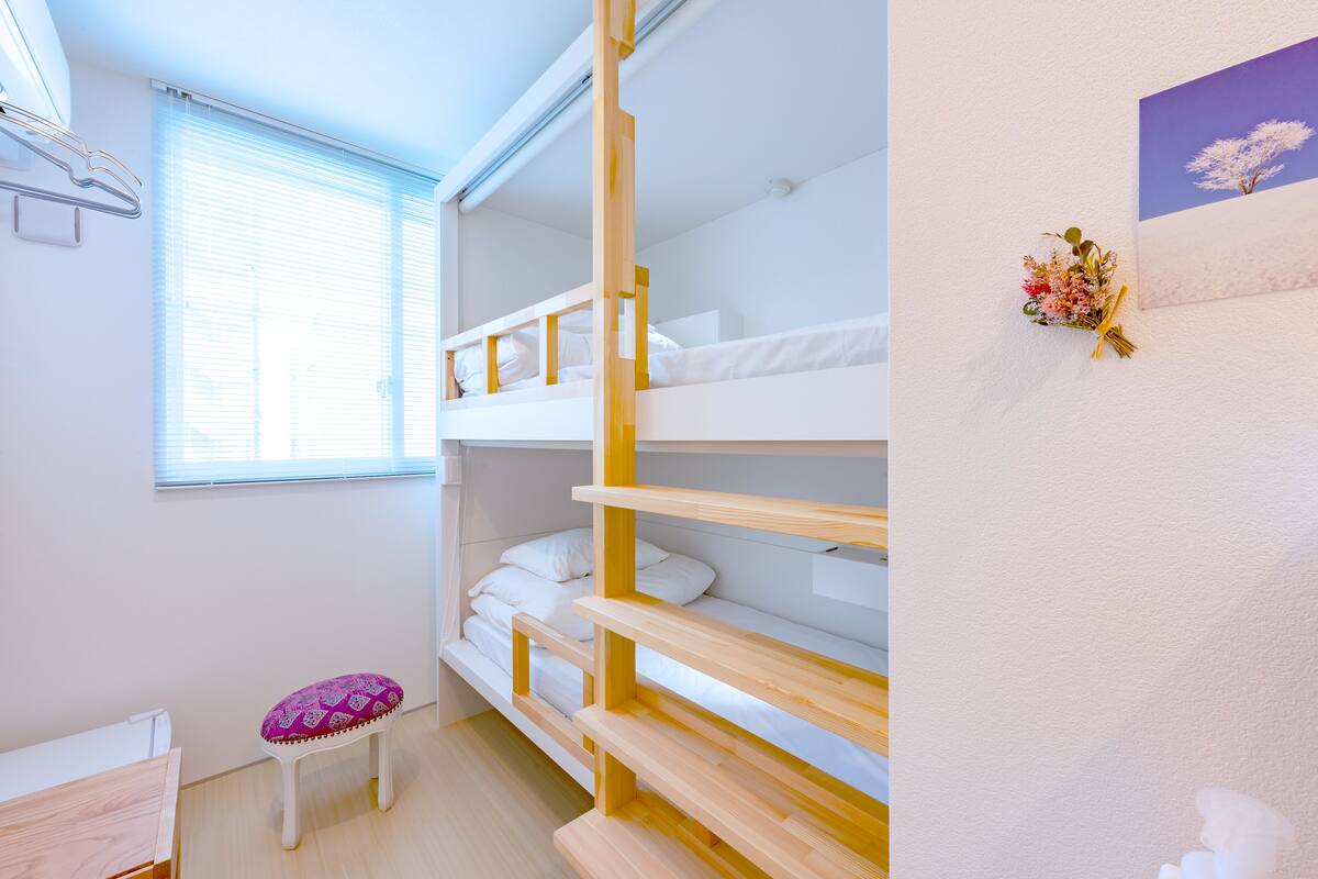 A bunk bed room with a small stool and a window with blinds.