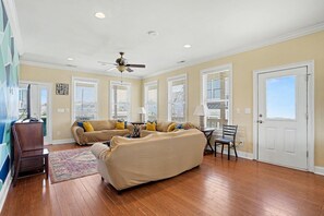 A spacious living room with a ceiling fan, large windows with blinds, a sofa set, a coffee table, and a television.