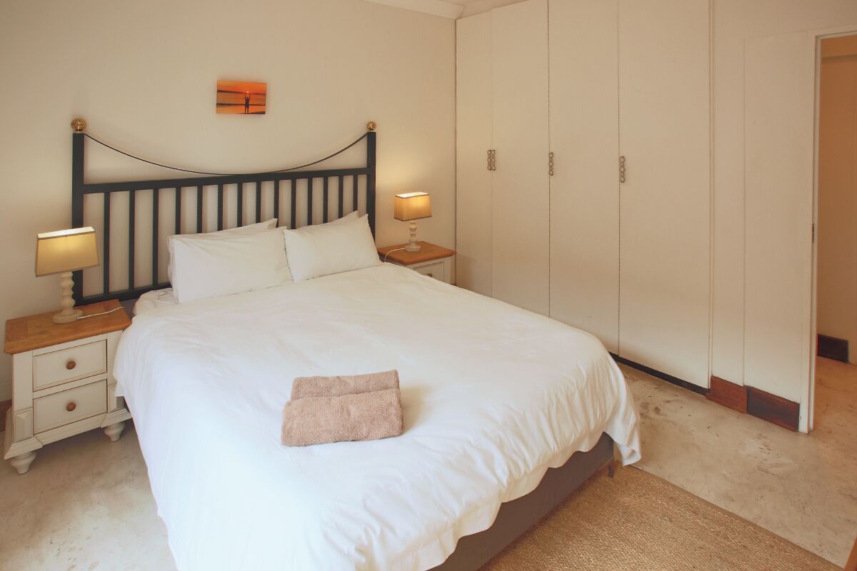 A neatly made bed with white linens, two bedside lamps, a wooden nightstand, a small drawer, and a single towel on the bed.