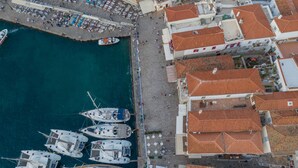 Vista aérea de un pueblo costero con un puerto deportivo, edificios con tejados de terracota y un paseo marítimo con sillas de playa y sombrillas.