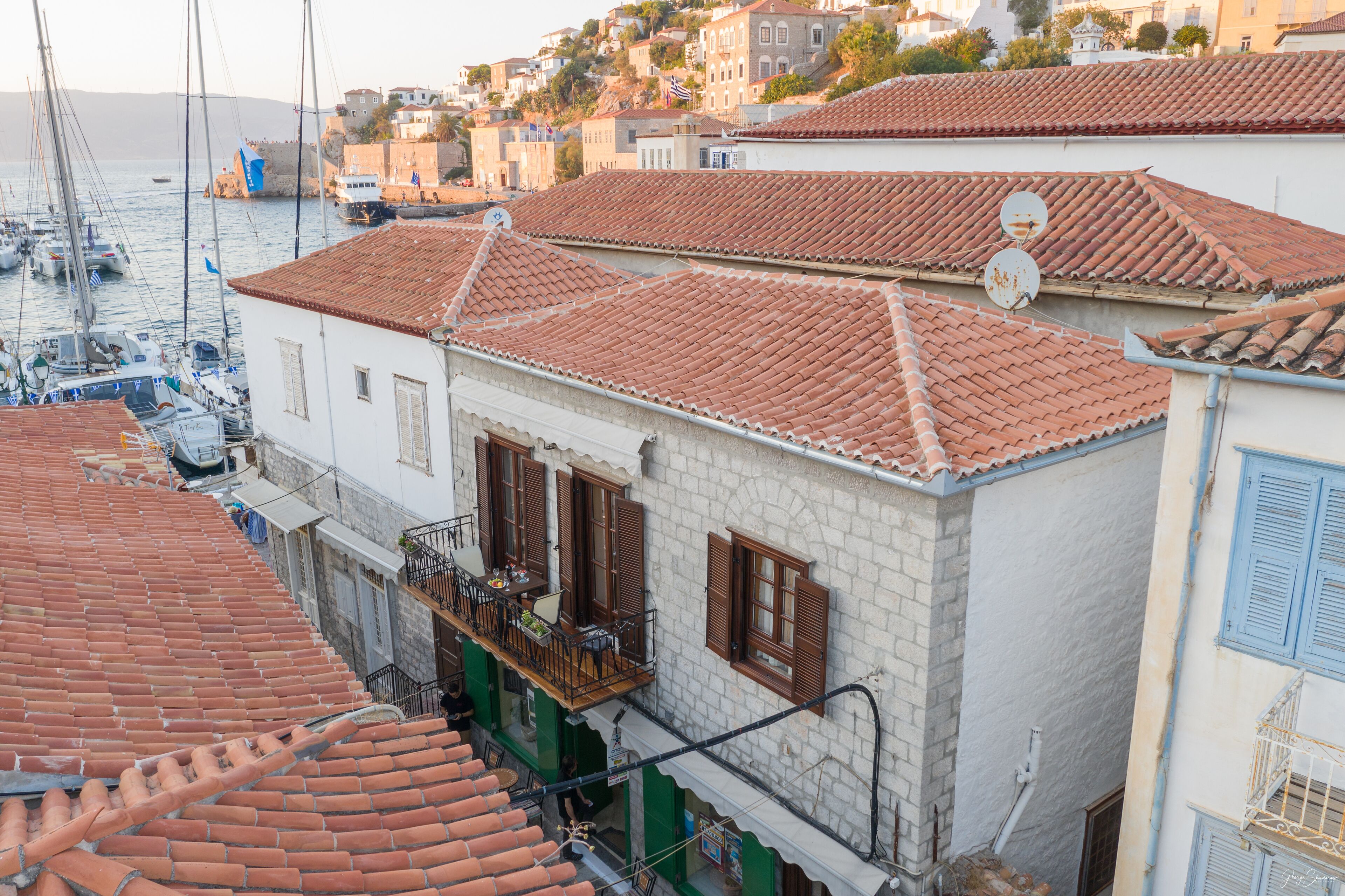 A coastal town with terracotta-tiled buildings, a balcony with outdoor furniture, and a view of boats in the water.