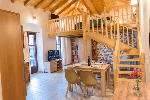 A cozy room with a wooden staircase, a dining table with chairs, a television, and a bookshelf. - Guarda Il Porto Apartment - Hydra port (Idra)
