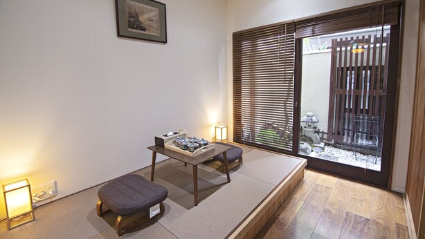 A traditional Japanese room with tatami flooring, a low table with cushions, a sliding door with a view of a garden, and a framed picture on the wall.