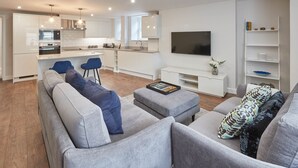 A modern living room with a grey sofa, ottoman, and blue chairs. A kitchen with white cabinets and a TV is visible in the background. - The Garden Apartment (Harrogate)