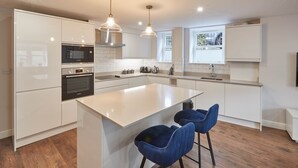 A modern kitchen with white cabinetry, a central island, two blue bar stools, and pendant lighting. - The Garden Apartment (Harrogate)