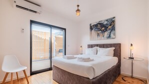 Une chambre avec un grand lit, une chaise blanche, une petite table et un tableau accroché au mur.