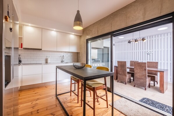 A modern kitchen with white cabinets, a wooden dining table, and a glass door leading to a dining area with wicker chairs.