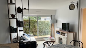 A compact living space with a bed, a desk, a chair, a bookshelf, a television, and a view of the outdoors through a sliding door. - Cabourg Mon Amour (Cabourg)