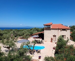 Une villa en pierre avec un toit recouvert de tuiles rouges, une piscine, du mobilier d’extérieur et une vue sur la mer.