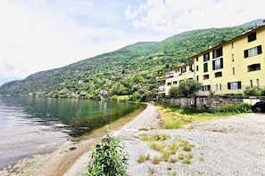 A lakeside building with a gravel path leading to the water, surrounded by greenery and hills. - Apartment on the beach facing Lake Como. Air conditioning, wifi, internal parking and beach access are free!The house is located right in front of the lake, from where you can enjoy the beautiful view of the lake. Bellano center and Bellano Tartavalle Te (Bellano)