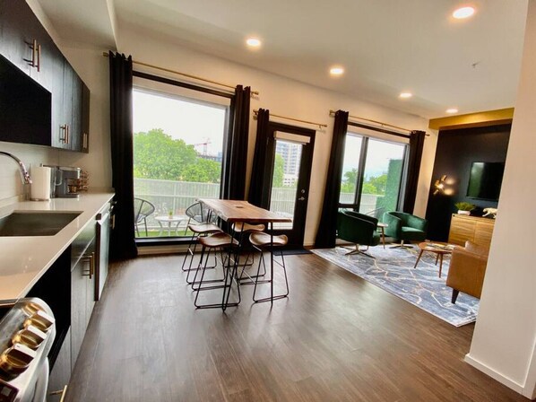 A modern kitchen with a dining area, large windows, and a view of greenery.