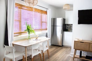 A modern kitchen with a stainless steel refrigerator, white cabinets, a dining table with chairs, and a large window with bamboo blinds. - The Beach Bungalow (Satellite Beach)