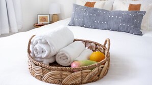 Un lit bien fait, avec du linge de lit blanc, un oreiller gris à pois blancs et un panier en osier contenant des serviettes enroulées et des fruits.