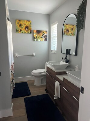 Een moderne badkamer met toilet, wastafel en spiegel. Twee aan de muur bevestigde schilderijen van zonnebloemen.