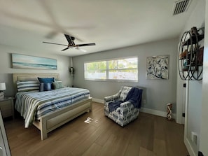 Une chambre à coucher avec un lit, une chaise, un ventilateur de plafond, une fenêtre et des œuvres d’art accrochées aux murs.
