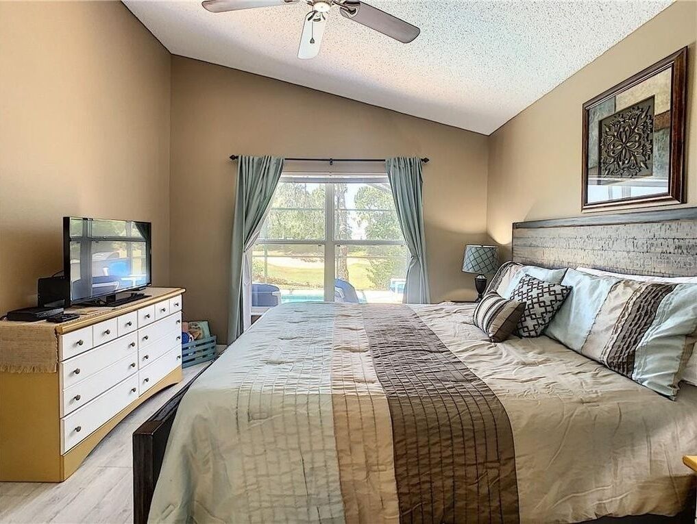 A bedroom with a bed, a dresser, a television, a ceiling fan, and a window with curtains.