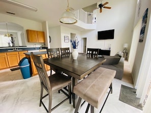 A dining area with a large table, chairs, and benches. There is a ceiling fan, a TV mounted on the wall, and a staircase leading to an upper level.