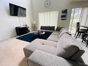 A living room with a sectional sofa, a flat-screen TV, a glass coffee table, and a dining area with a chair.