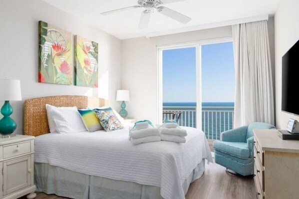 A hotel room with a bed, a desk, a chair, and a large window with a view of the sea.
