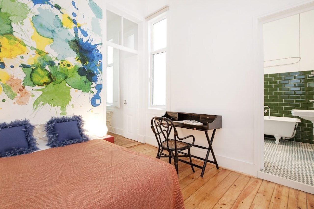 A bedroom with a bed, a desk, a chair, a window, and a bathroom with a bathtub and green tiles.