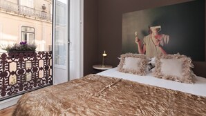Un dormitorio con un ventanal grande, un balcón con barandilla decorativa, una cama con manta marrón, una mesita de noche con lámpara y un cuadro en la pared.