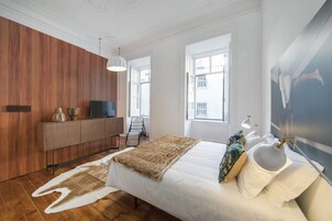 Un dormitorio moderno con un panel de madera en la pared, una cama con edredón blanco, una manta de piel y un ventanal con vistas a edificios.