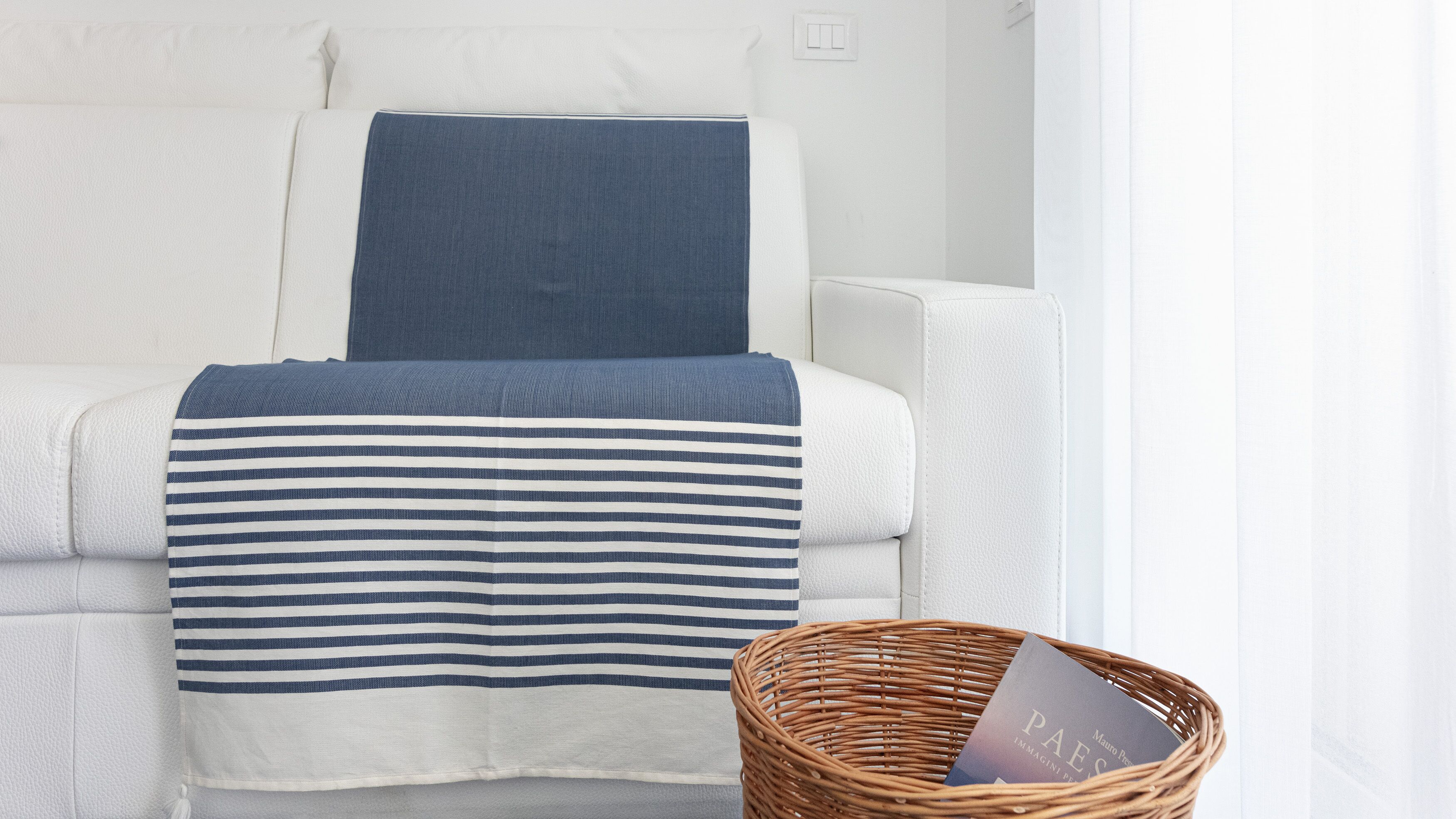 Un canapé blanc, agrémenté d’un plaid rayé bleu et blanc et d’un coussin bleu foncé. Un panier en osier contenant un livre est posé sur le sol, à côté du canapé.