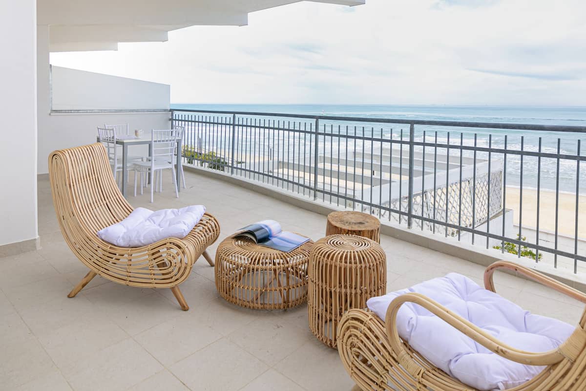 A balcony with wicker furniture, a table, and chairs, overlooking a beach and the ocean.