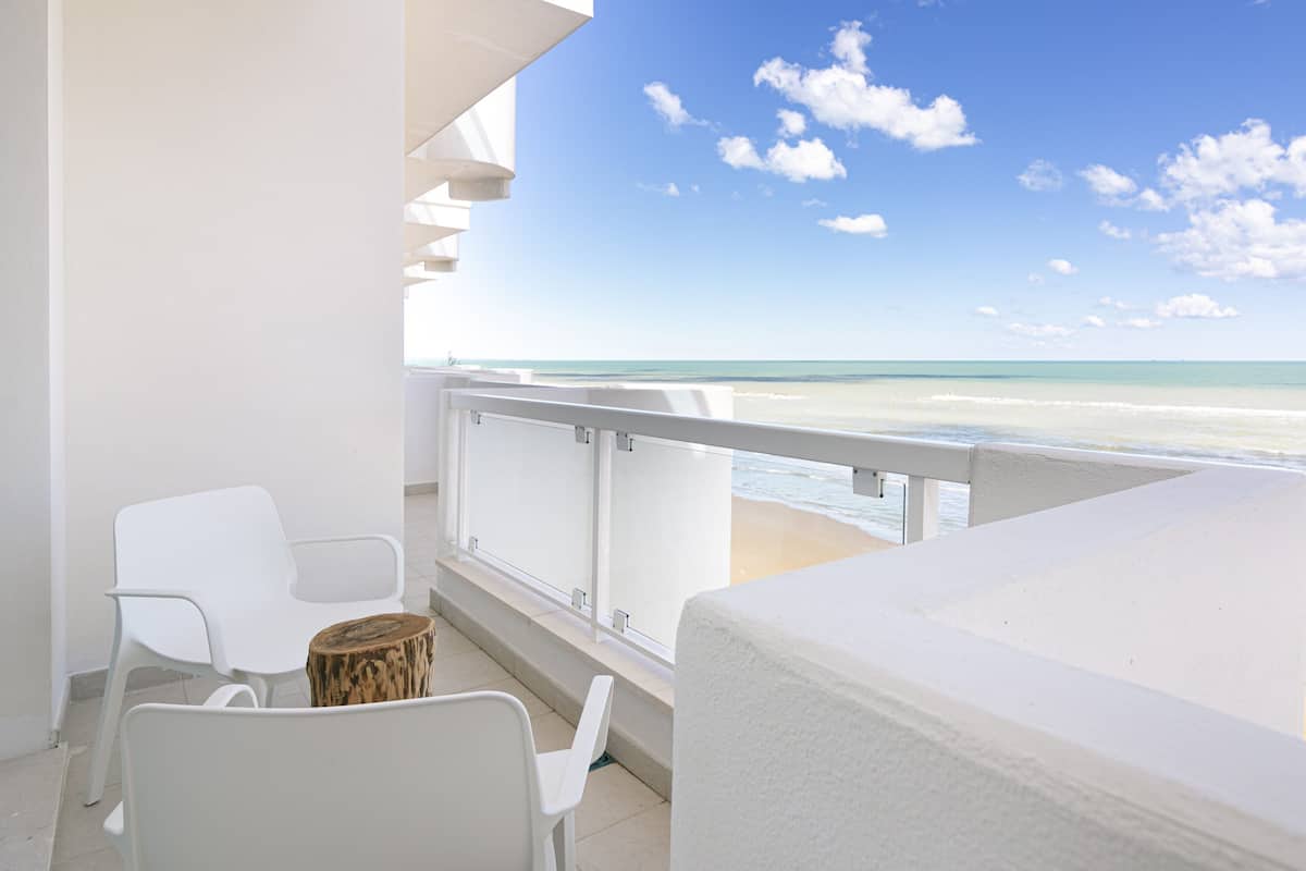 A balcony with white chairs and a wooden stool, overlooking a beach with clear skies and calm waters.