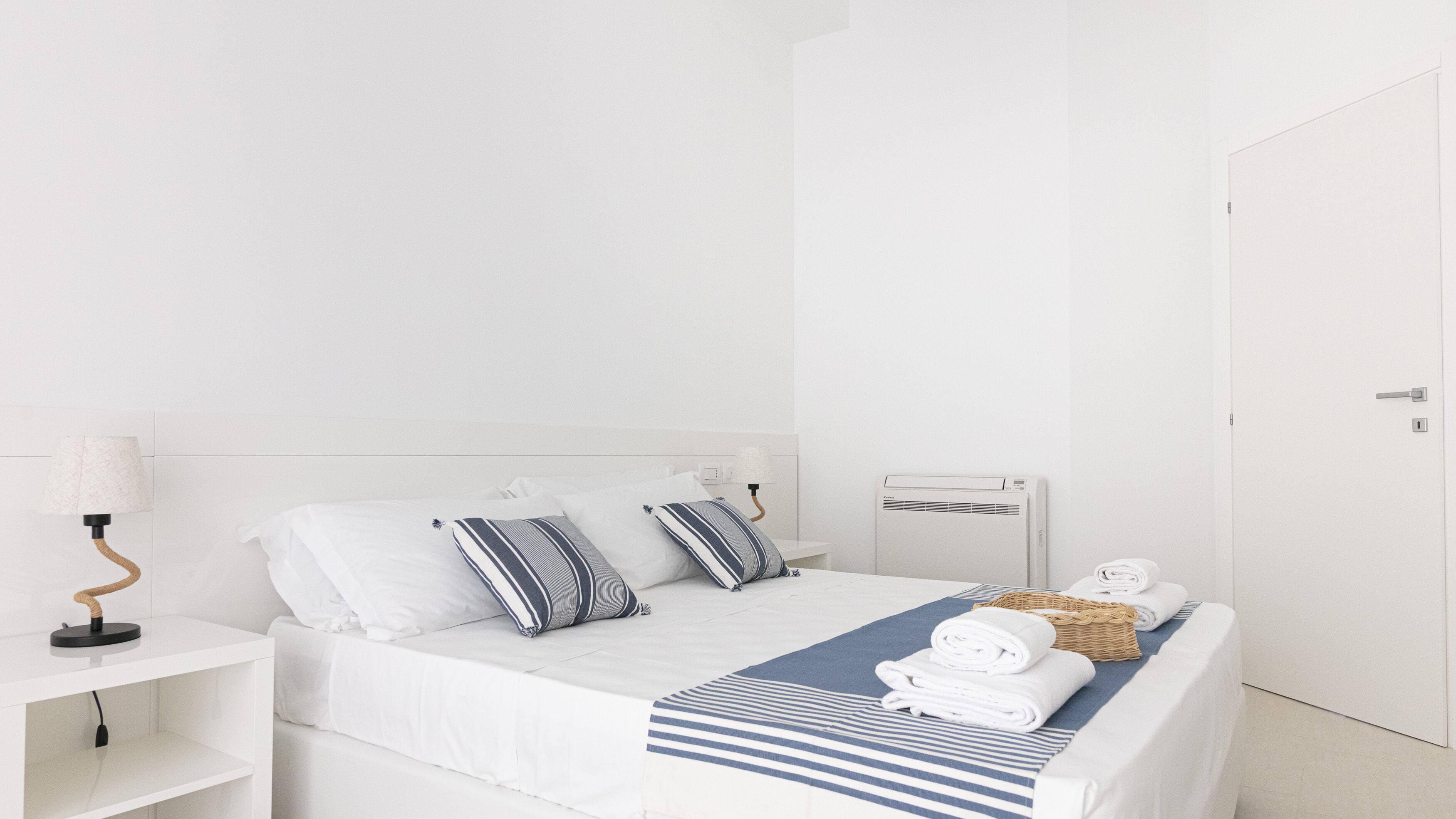 A minimalist white hotel room with a bed, a bedside table with a lamp, a wall-mounted air conditioner, and a door.