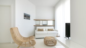 Un salon moderne avec un canapé blanc, un fauteuil en osier, un pouf rond, un téléviseur à écran plat et un tableau encadré au mur.