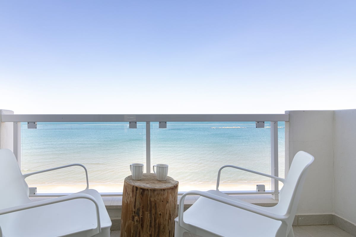 A balcony with two white chairs and a small table, overlooking a beach with clear blue water.