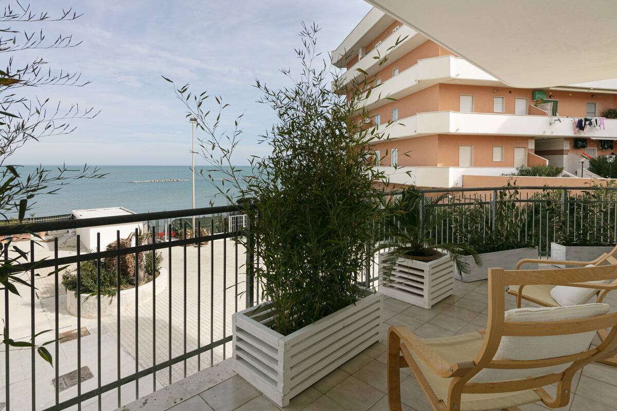 A balcony with a view of the sea, a wooden chair, and a potted plant.