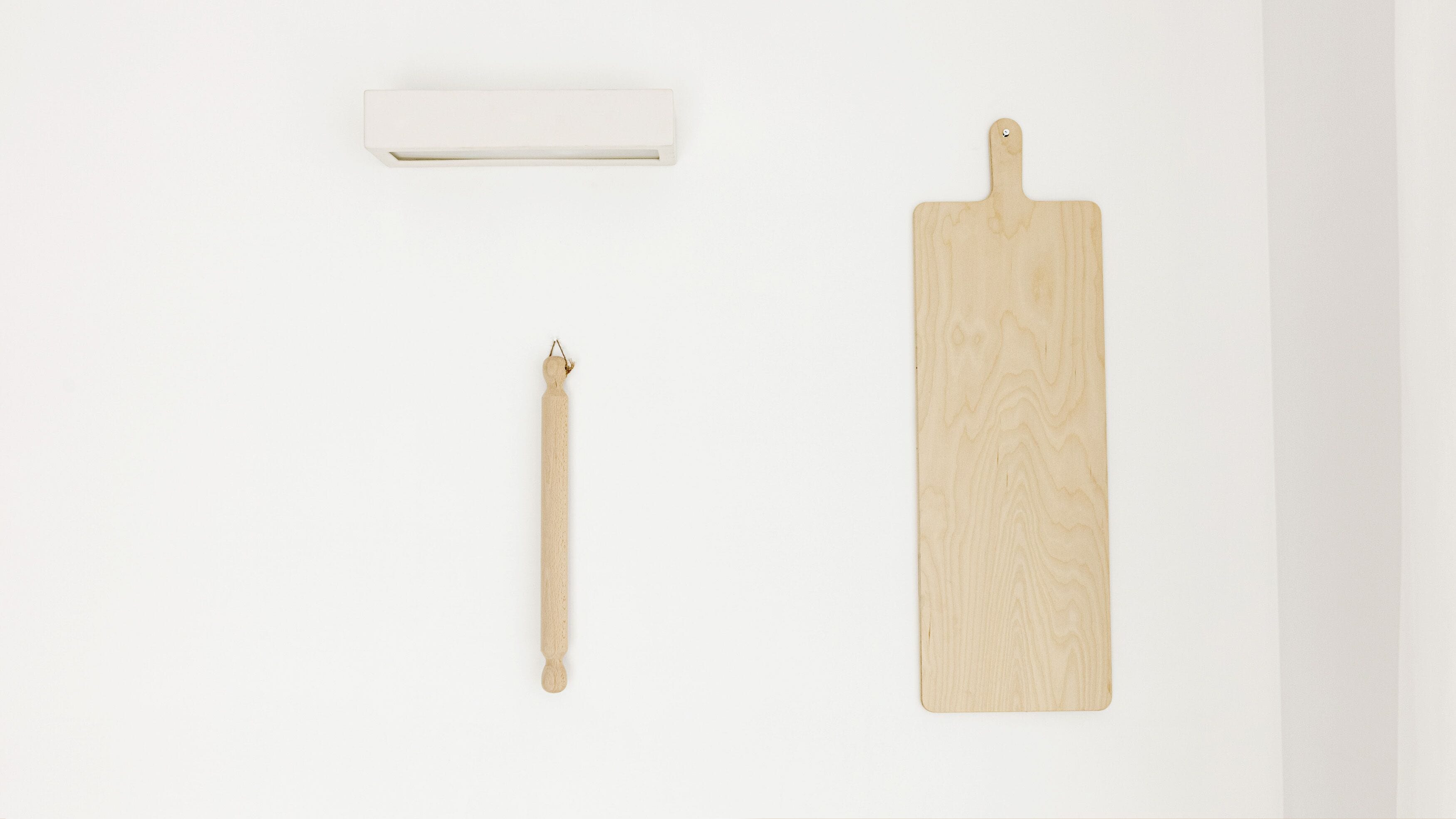 A wooden cutting board, a wooden spoon, and a white air vent on a white wall.