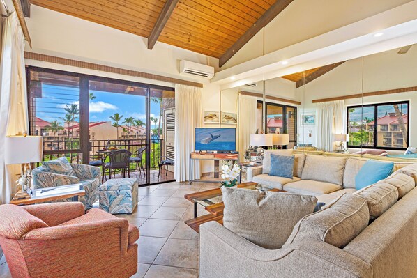 A spacious living area with a large sofa, a coffee table, and a TV. There is a balcony with a view of palm trees and buildings.