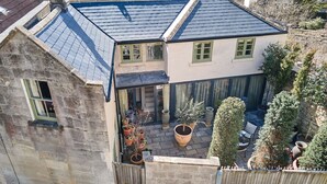 A two-story house with a sloped roof, a stone wall, and a small patio with potted plants and outdoor furniture. - Oppulent Lansdown Crescent Mews House with Free parking - Pass the Keys (Bath)