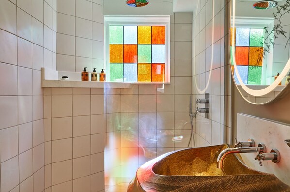 A bathroom with a large, ornate sink, a window with colorful stained glass, and a mirror with a plant in the reflection.