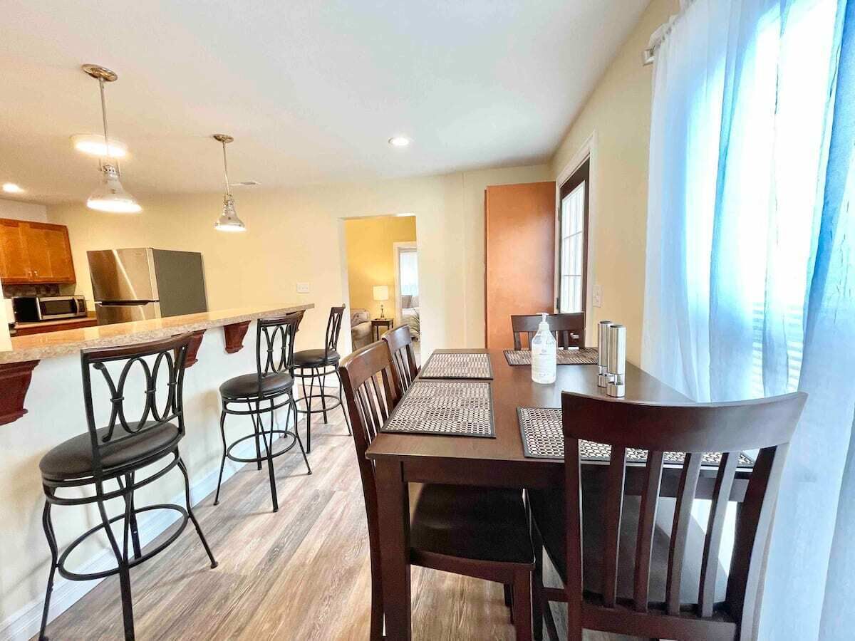 A dining area with a wooden table and chairs, a kitchen in the background, and a window with curtains.