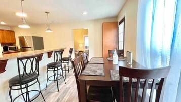 A dining area with a wooden table and chairs, a kitchen in the background, and a window with curtains.
