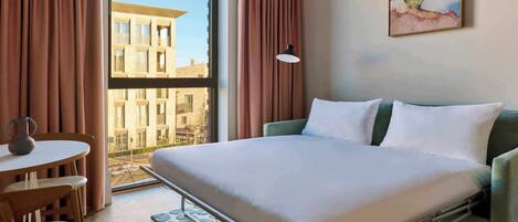A hotel room with a bed, a table with chairs, a window with curtains, and a painting on the wall.