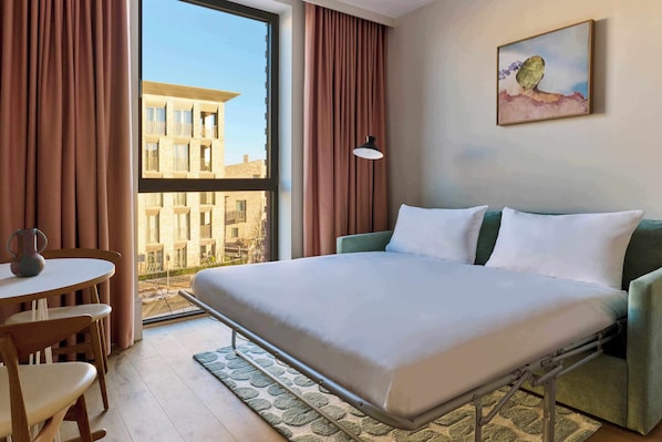 Habitación de hotel con cama, una mesa con sillas, una ventana con cortinas y un cuadro en la pared.