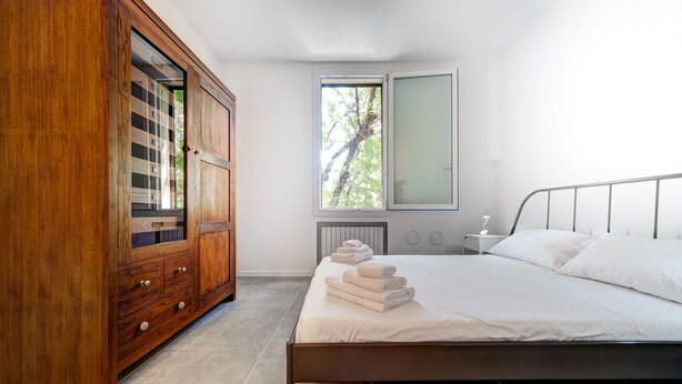 A bedroom with a bed, a wooden wardrobe, a window, and a bedside table with a lamp.