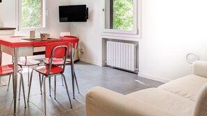 A modern room with a red dining table, four chairs, a beige sofa, a flat-screen TV mounted on the wall, and large windows with a view of greenery.