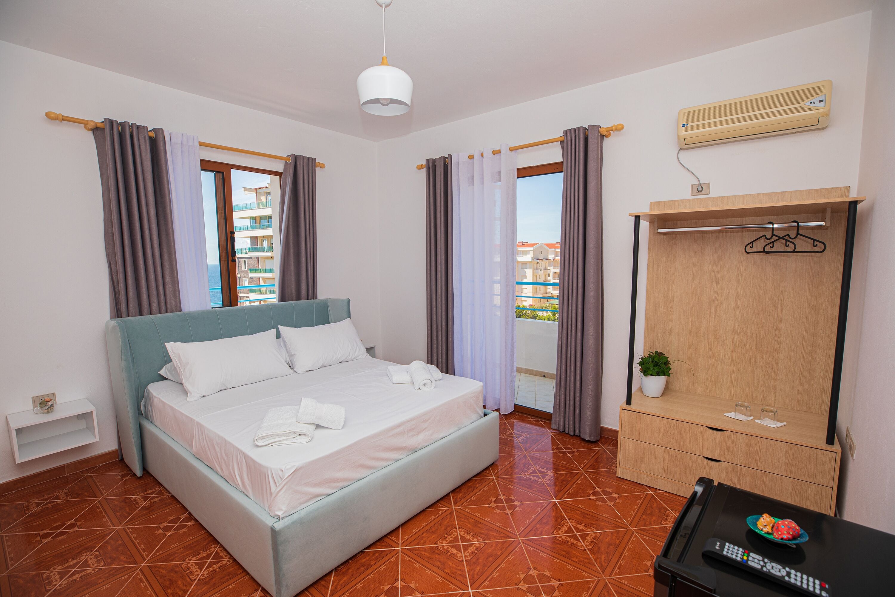 A bedroom with a bed, a wardrobe, a small table with a remote control, and a potted plant.