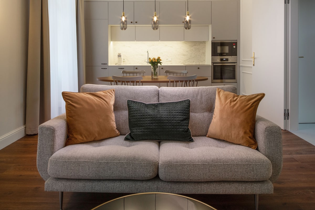 A modern living room with a grey sofa, two brown cushions, and a dark green accent pillow. In the background, there is a kitchen with white cabinets, a marble backsplash, and stainless steel appliances.