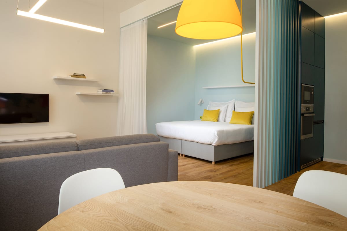A modern hotel room with a sofa, a bed with yellow pillows, a wooden table, and a TV on the wall.