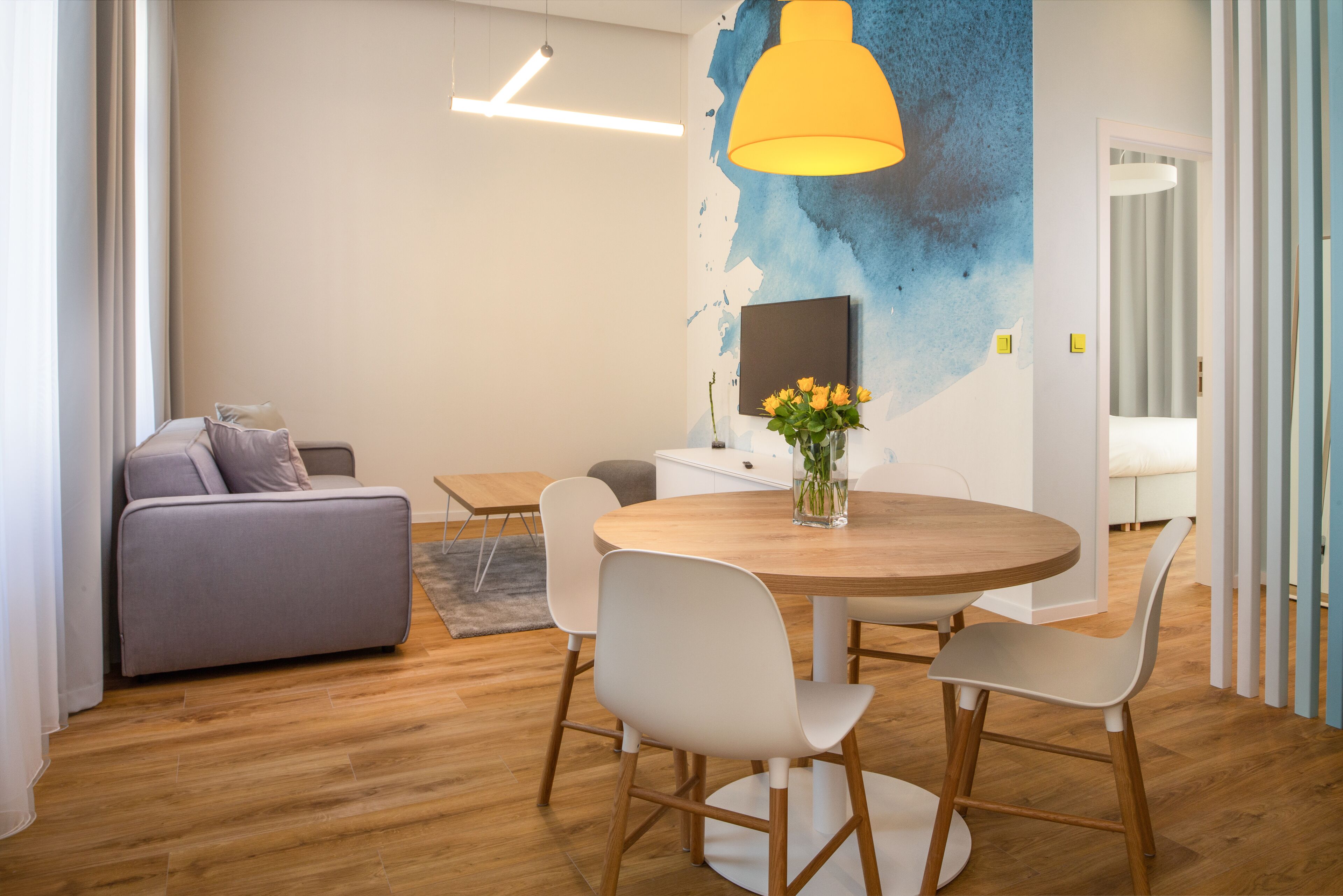 A modern living room with a round wooden table, white chairs, a grey sofa, and a yellow pendant light.