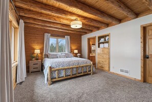 Un dormitorio con techo de madera, una cama con ropa de cama estampada, una mesita de noche, una ventana con cortinas y un armario de madera.