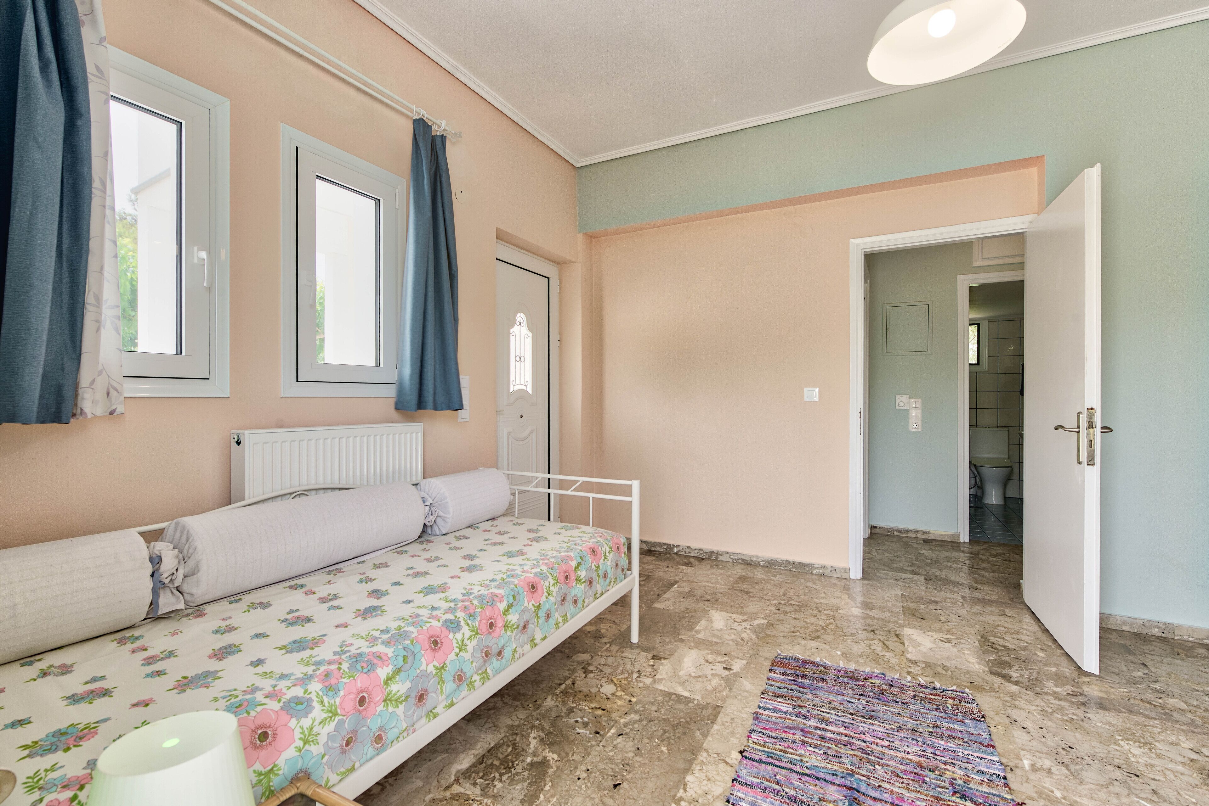 Quarto com cama, radiador, janela com cortinas, porta e banheiro visível através de uma porta aberta.