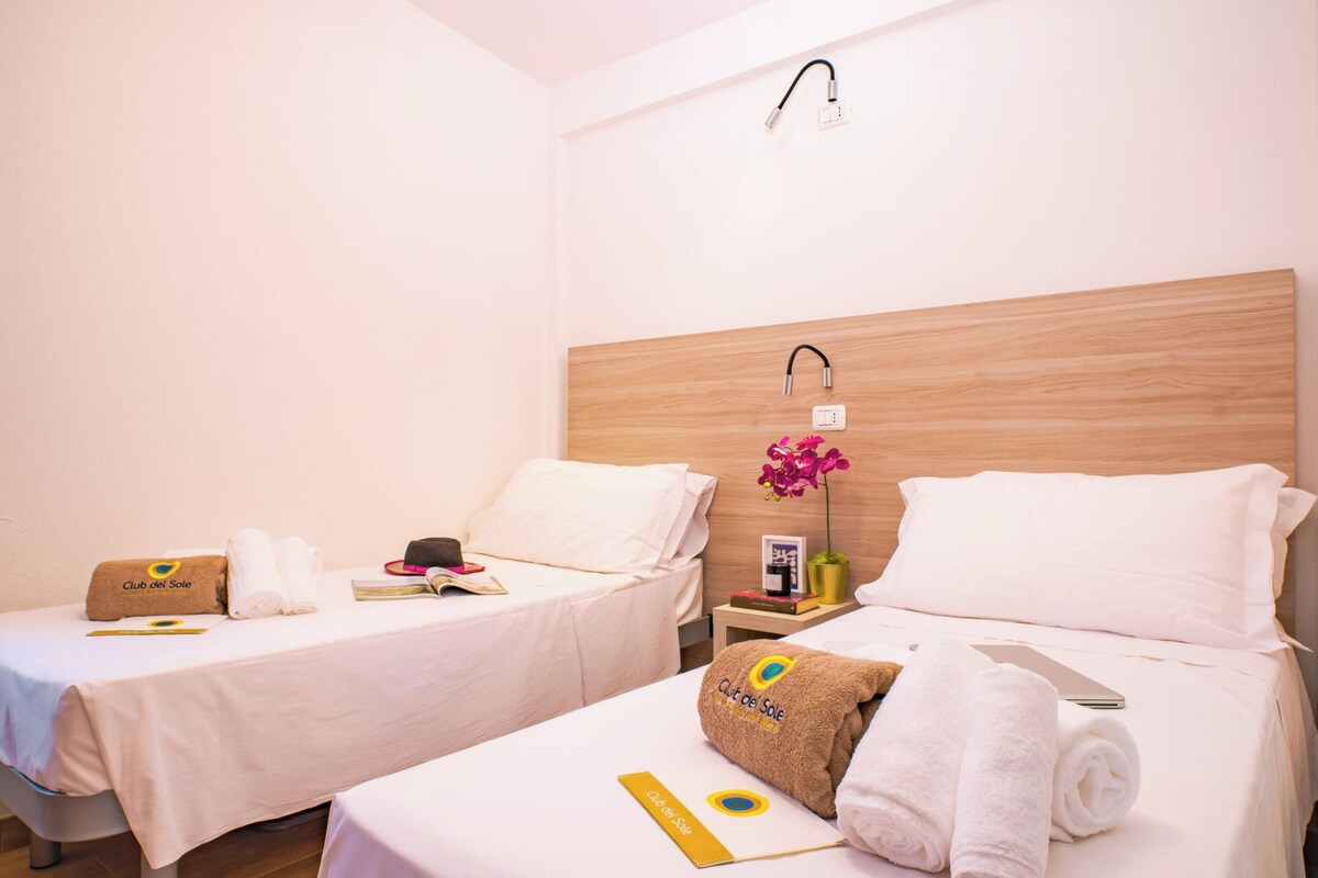 A hotel room with two beds, each with a white bedspread and a towel with the hotel's logo. There is a wooden headboard on one bed and a nightstand with a lamp and a small potted plant.